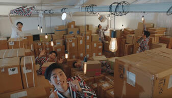 Multiple versions of the same person wearing the same outfit are popping out amongst packing boxes that are filling a room